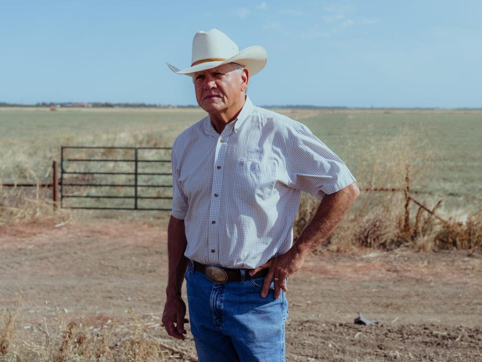 scott stone rancher in jeans button down shirt and cowboy hat stands in dirt lot next to field of grass