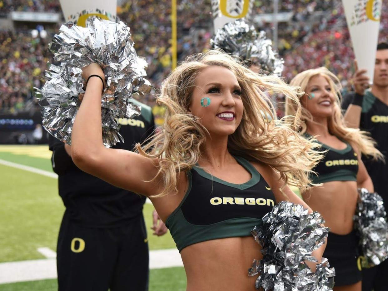 Chloe is dressed in a cheering uniform that says "Oregon" on the top. She holds silver pom poms, and both male and female cheerleaders cheer behind her.