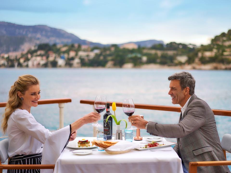 A couple raising their wine glasses to one another while sitting down for a meal on the deck of The World.