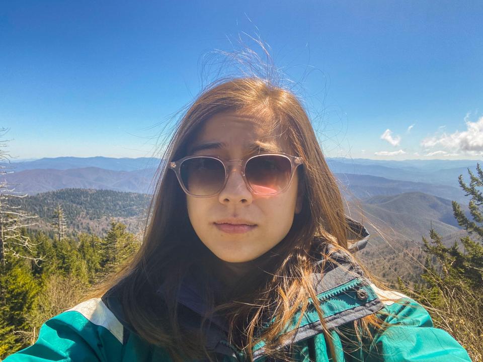 The author wearing sunglasses and a teal jacket with trees, mountains, and a clear blue sky behind her