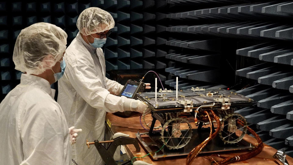 two scientists stand next to one of the rovers that's been placed on a table in a special chamber with lined walls