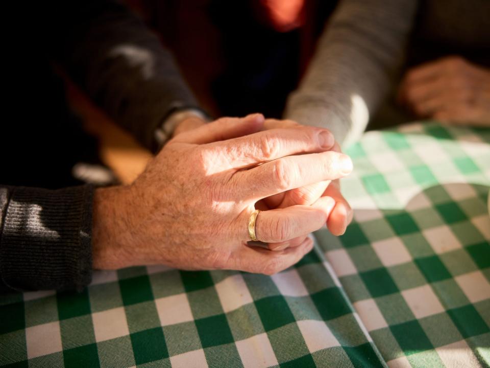 Hands of an elderly person, wearing a gold ring, holding another person's hand in warm sunlight over a cafe table.