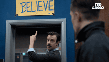 Ted Lasso points to his "Believe" sign