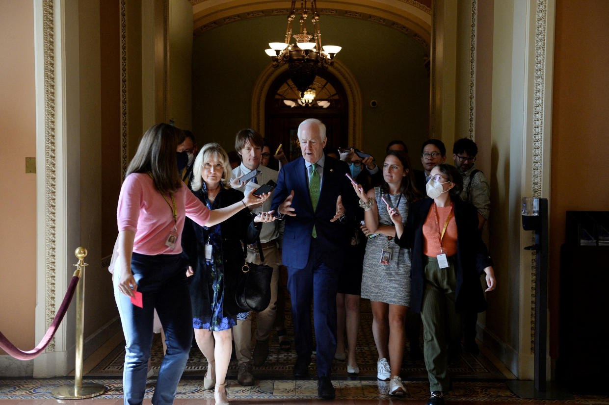 Sen. John Cornyn, walking down a corridor at the Capitol, answers questions as reporters surround him.