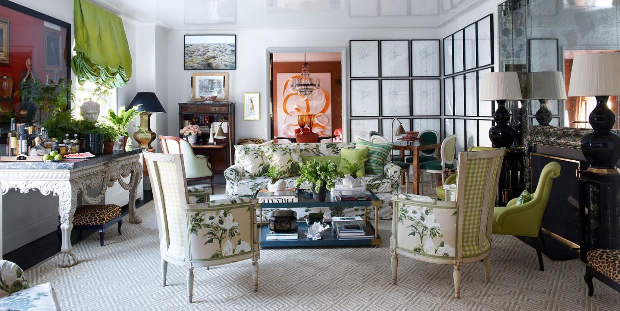 large living room with bright white lacquer walls and ceiling and two girds of nine frames in the far right corner and a sofa and upholstered straight back chairs at center and a two tier cocktail table and on the side is a large claw foot table with low bench underneath and a console with matching black lamps on either end on the right wall