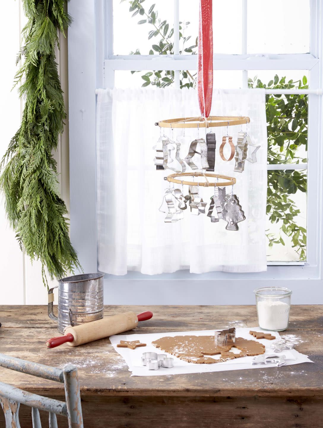 a mobile made from embroidery hoops and cookies cutter haning in front of a window, and above a table that has gingerbread cookie dough on it