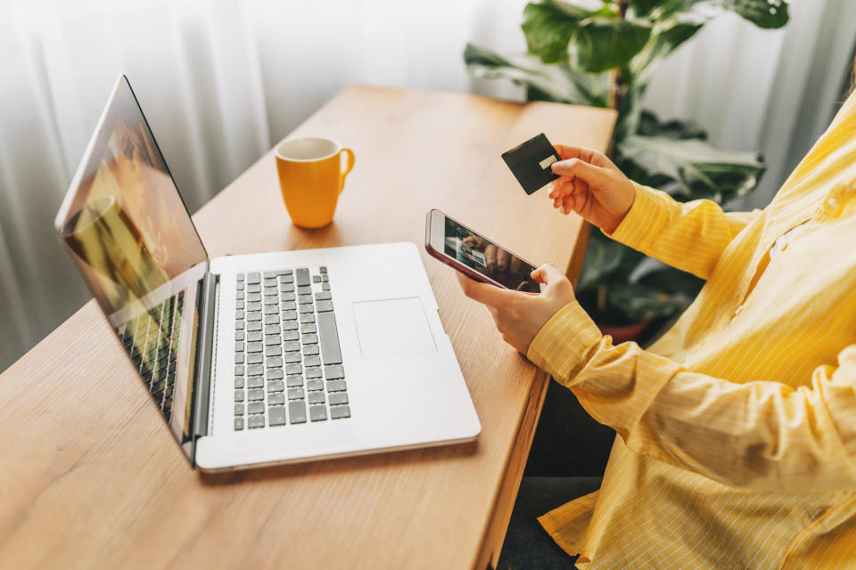 Person in a yellow shirt holds a smartphone and a credit card, sitting at a desk with a laptop, mug, and plant