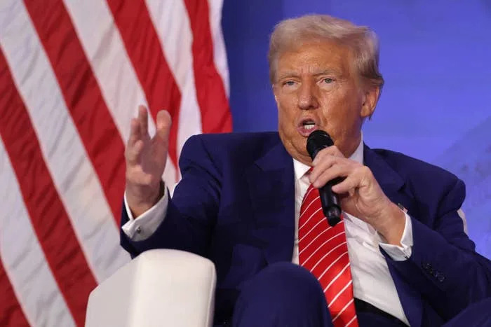 Donald Trump speaking into a microphone, gesturing with his hand, seated in front of a U.S. flag