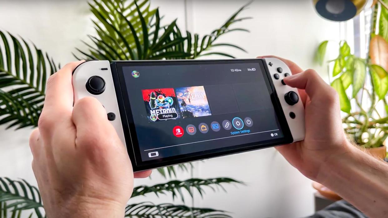  A pair of hands holding a Nintendo Switch showing the System Settings icon highlighted on the home screen. 