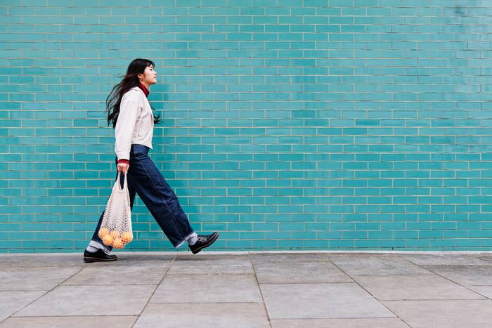 A person is walking alongside a turquoise brick wall, holding a netted bag. They are wearing a light jacket, red shirt, and wide-leg jeans