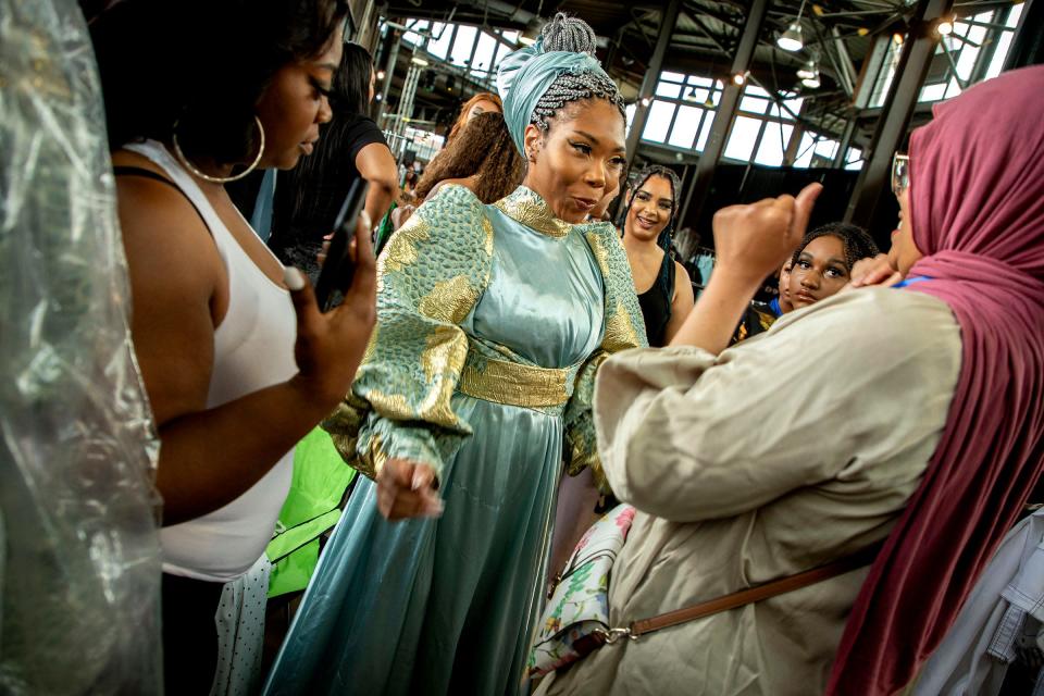 Designer LaTerry Moshin, of Sew Modest Studio, right, smiles and claps reacting after she put the finishing touches on the outfit she designed, which is being worn by model Fai Marie, center as they prepare to walk the runway for the Michigan Fashion Week's 10th anniversary show at Eastern Market on June 10, 2022.