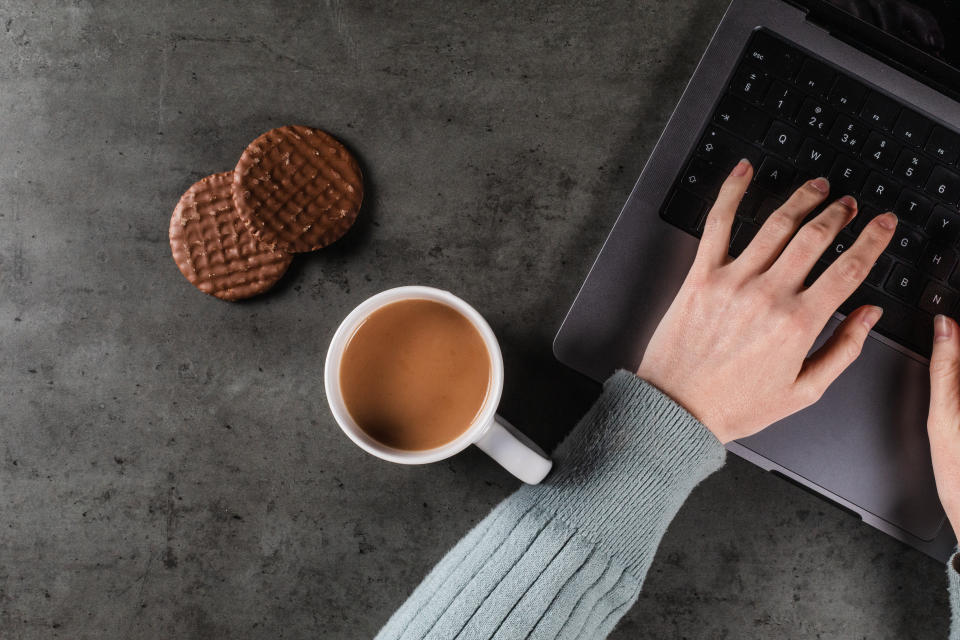 A caucasian person using a modern laptop computer with a mug of tea and chocolate biscuits on a dark textured work desk.