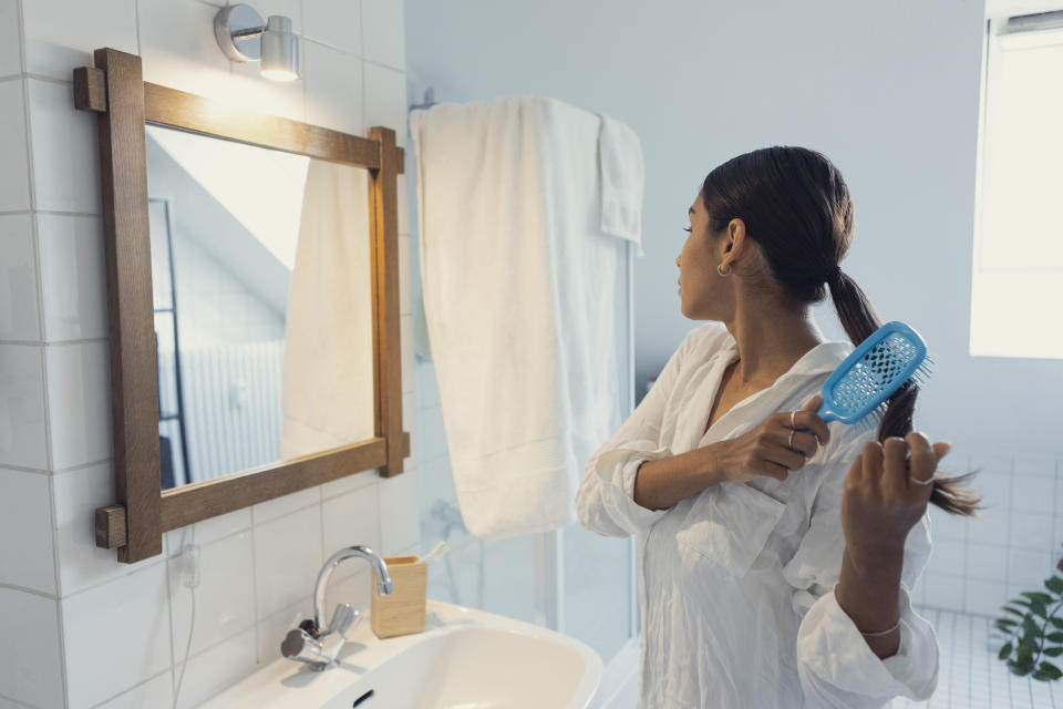 Woman in white pajamas brushing hair and looking in mirror at home