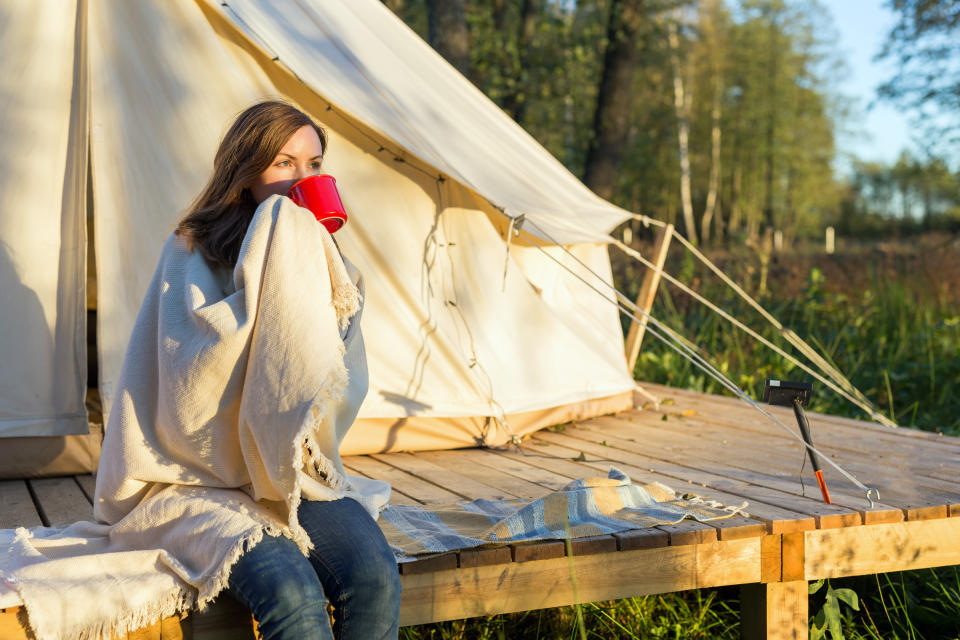 Young woman wraps blanket over herself while sitting and drinking coffee near canvas tent in the morning in the woods