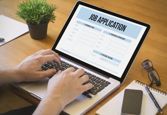 Two hands type on a laptop sitting on a wooden desk. An online job application form appears on the laptop screen.