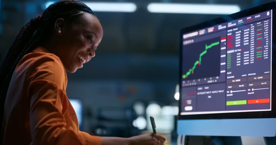 girl in orange sweater smiling in front of stock market screen and writing something down