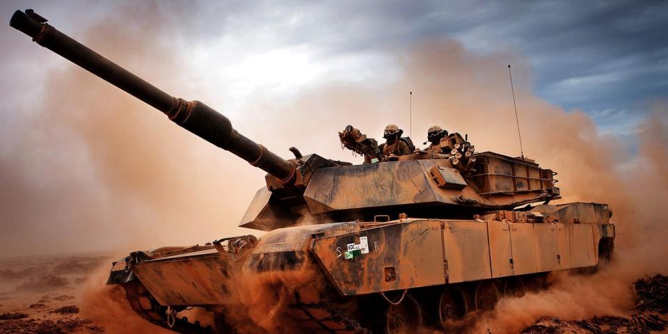 Marines in an M1A1 Main Battle Tank roll down a dirt road, kicking up dust behind them.