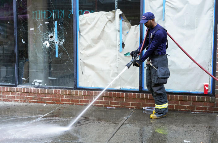 A fireman uses a hose to clean a sidewalk in front of a storefront in which bullet holes are visible.