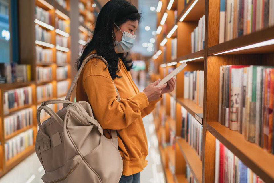 An Asian woman with a backpack stands by a library shelf reading a book