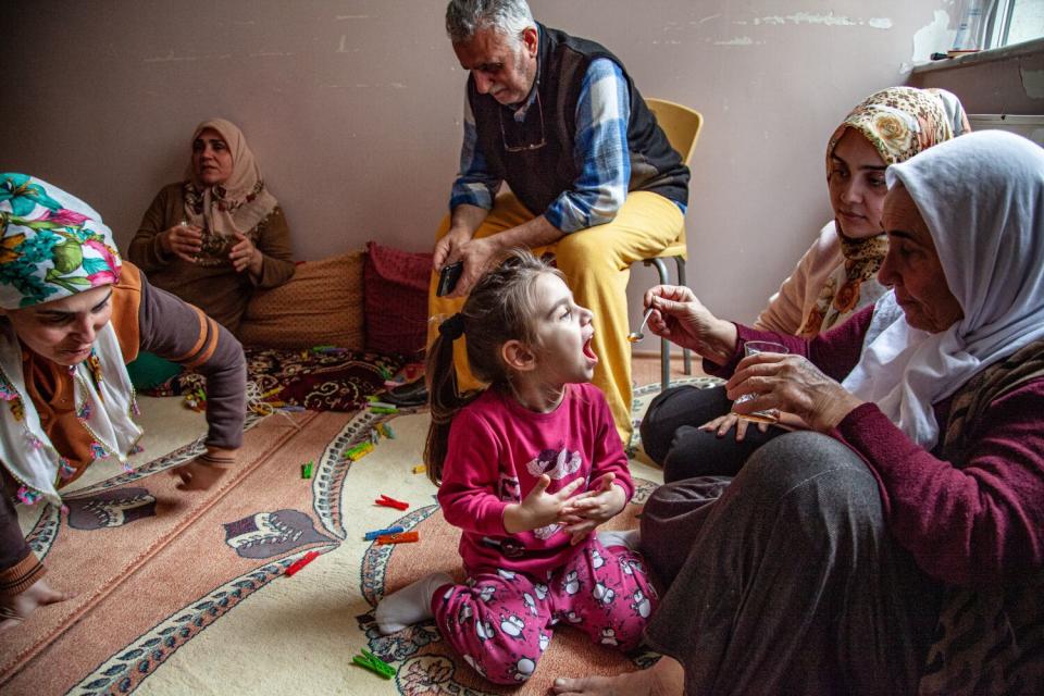 A man sits in a chair near four women in headscarves who are seated on a rug, one feeding a girl in pink clothes