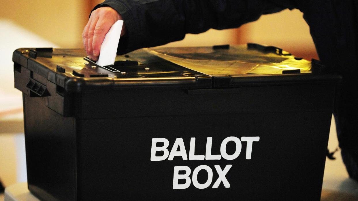 A hand is putting a piece of paper into a black box, that has the writing "ballot box" on the side in white letters