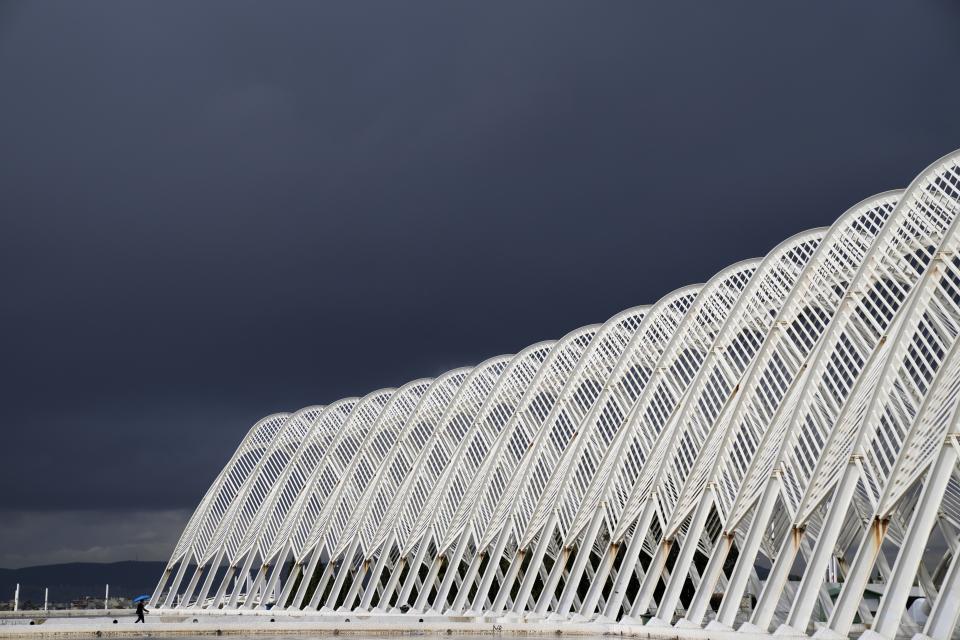 A man holding an umbrella walks at the modern Agora walkway at the main Olympic complex as a rainstorm approaches Athens, Thursday, Oct. 14, 2021. Greece's authorities are in full activation mode as the wet and stormy weather system named "Ballos" reached the country bringing thunder, lightning and showers, and causing fluctuations in the power supply. (AP Photo/Thanassis Stavrakis)