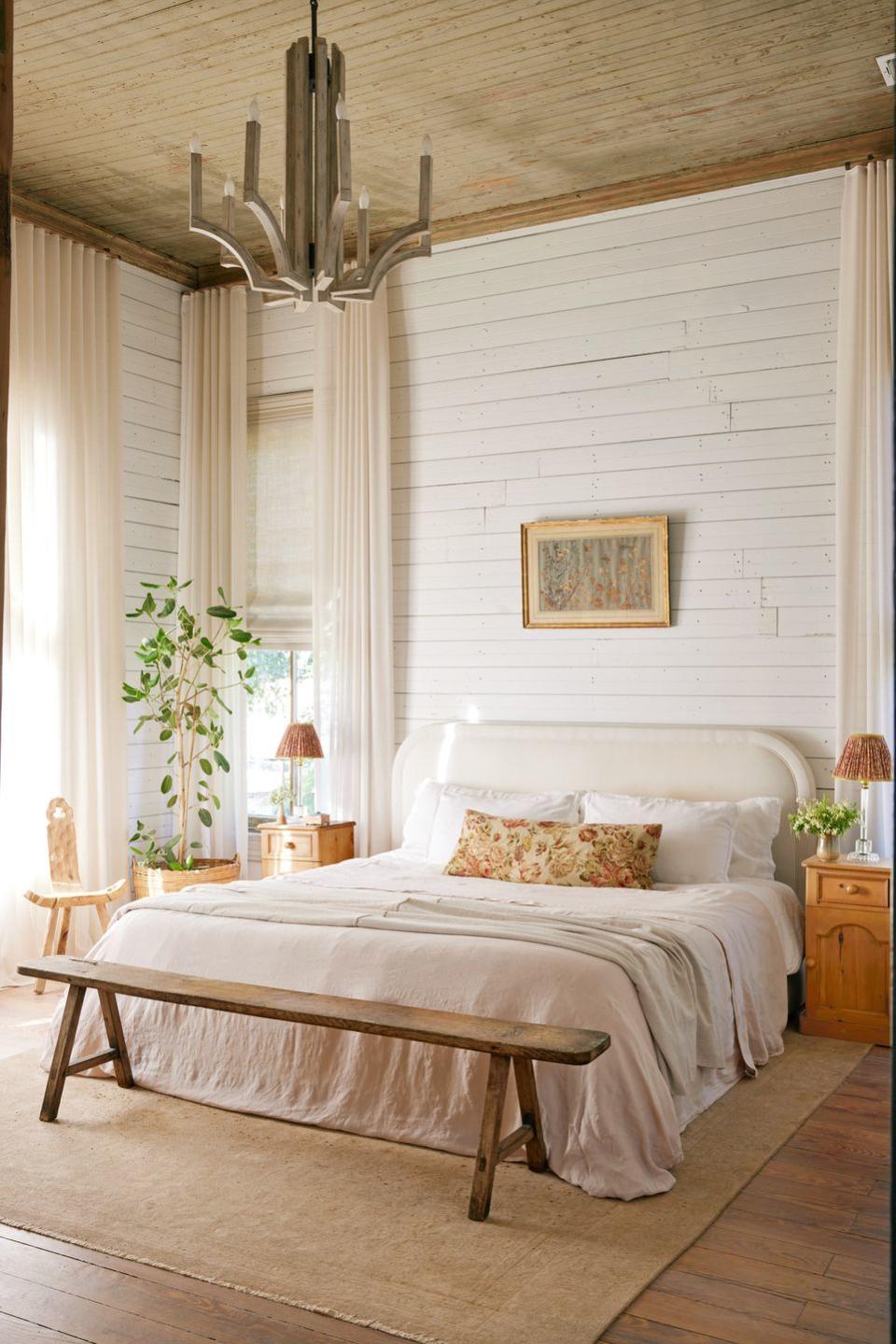 linen draperies hang in a bedroom against white shiplap walls and the bed is all white too with a floral pillow