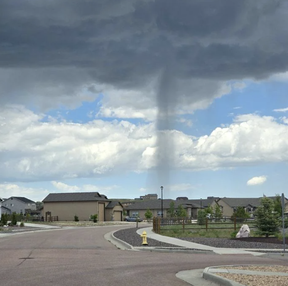 A funnel cloud forms over a suburban residential area. Houses and a fire hydrant can be seen in the foreground.