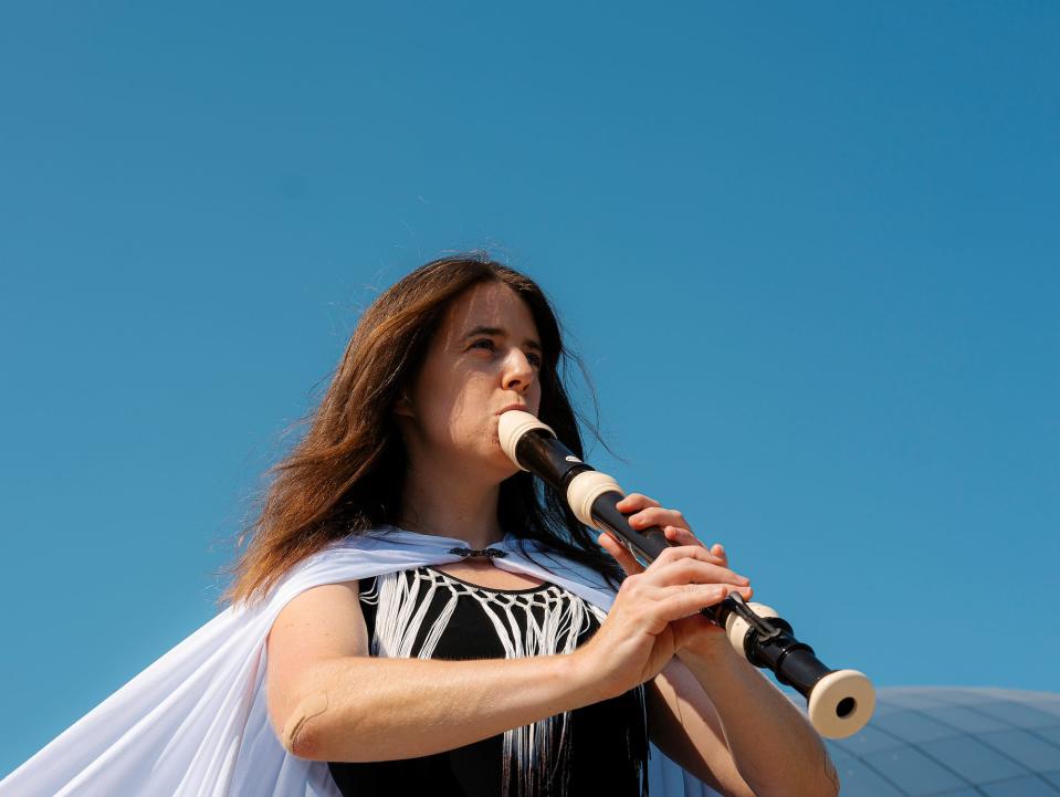 Liza Bec is wearing stage clothing outdoors playing a recording on a blue sky background