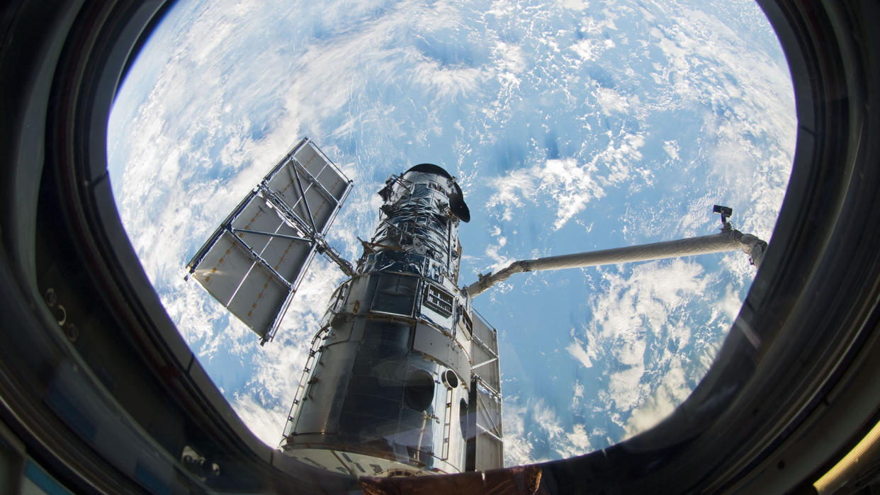  A view of a telescope and robotic arm through the window of a space shuttle. earth and clouds are in the background. 