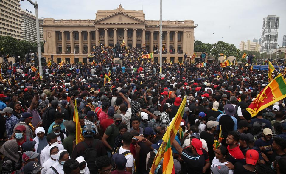 A large crowd of protestors waving flags gather in front of the presidential building in Sri Lanka.