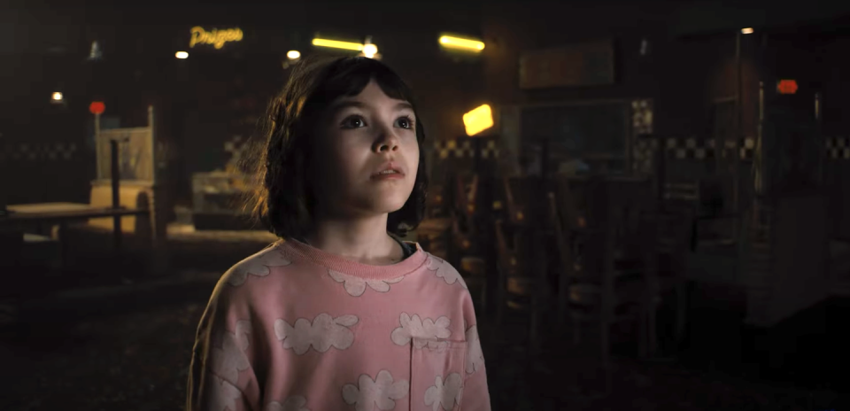 piper rubio in five nights at freddys, a young girl with dark hair in a bob, wearing a pink top with clouds on, she stands in a closed diner looking up at someone