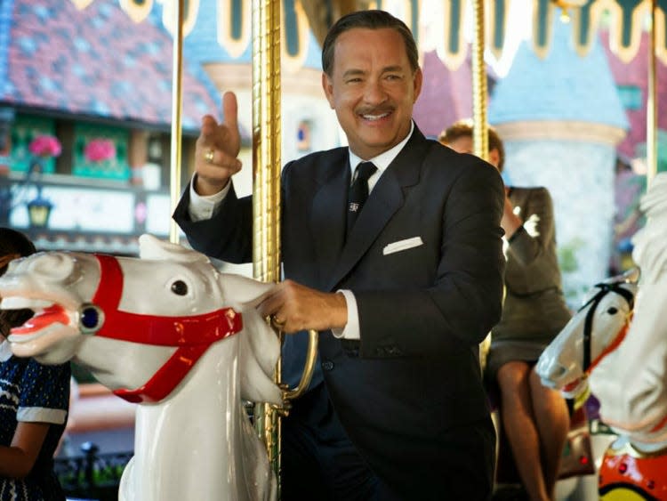 Tom Hanks dressed in a suit and with a mustache giving the two finger point while riding on a carousel.