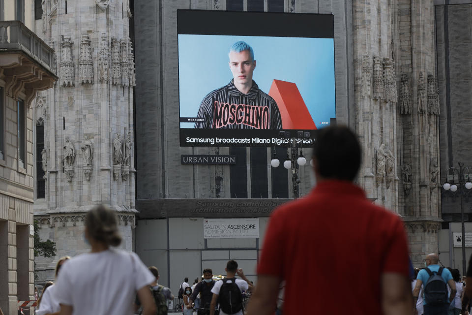 Peatones pasan por una pantalla en la catedral Duomo que muestra a un modelo de Moschino durante la Semana de la Moda Digital de Milán en Milán, Italia, el martes 14 de julio de 2020. La Semana de la Moda cambió a un formato digital por el COVID-19. (Foto AP/Luca Bruno)