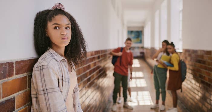 A young girl leaning against a wall as other kids laugh at her in the background