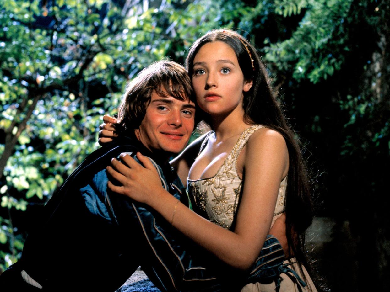 A teenage boy and girl dressed in period costumes embrace by a wall of green trees and bushes.