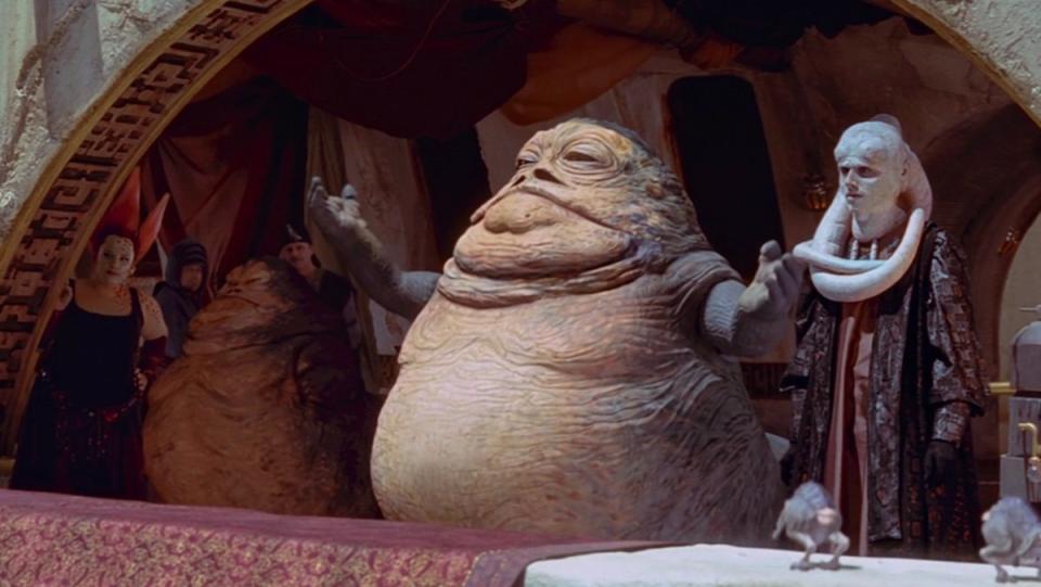 Jabba the Hutt walks out with his arm raised to greet the crowd at the Boonta Eve Classic in The Phantom Menace