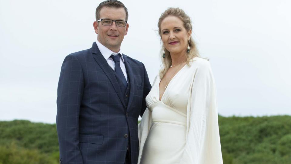 2021 MAFS couple Beth and Russell outside at their show wedding. He wears a dark blue suit and tie and has glasses. She wears a waisted gown with a loose cape. Her blonde hair is partially tied back.