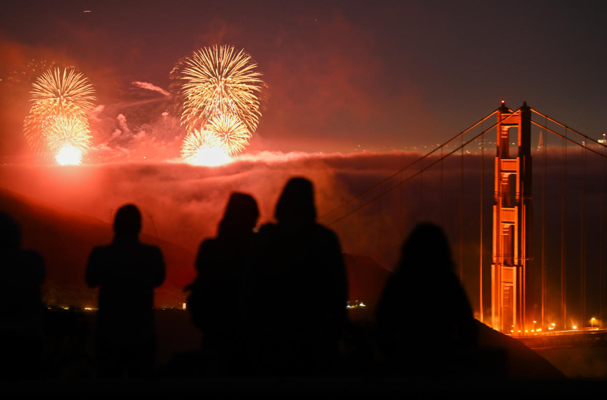 Silhouettes of people sitting together at Marin Headlands watching fireworks as they illuminate the sky over San Francisco's Golden Gate Bridge.