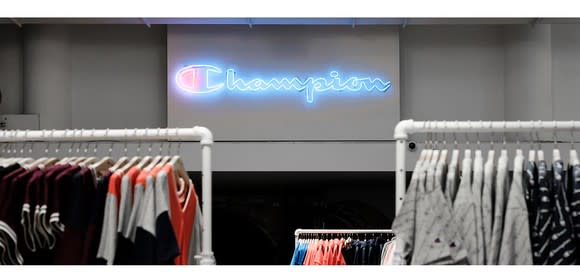 A brightly lit Champion logo on a grey wall inside a store with clothing racks in the foreground.