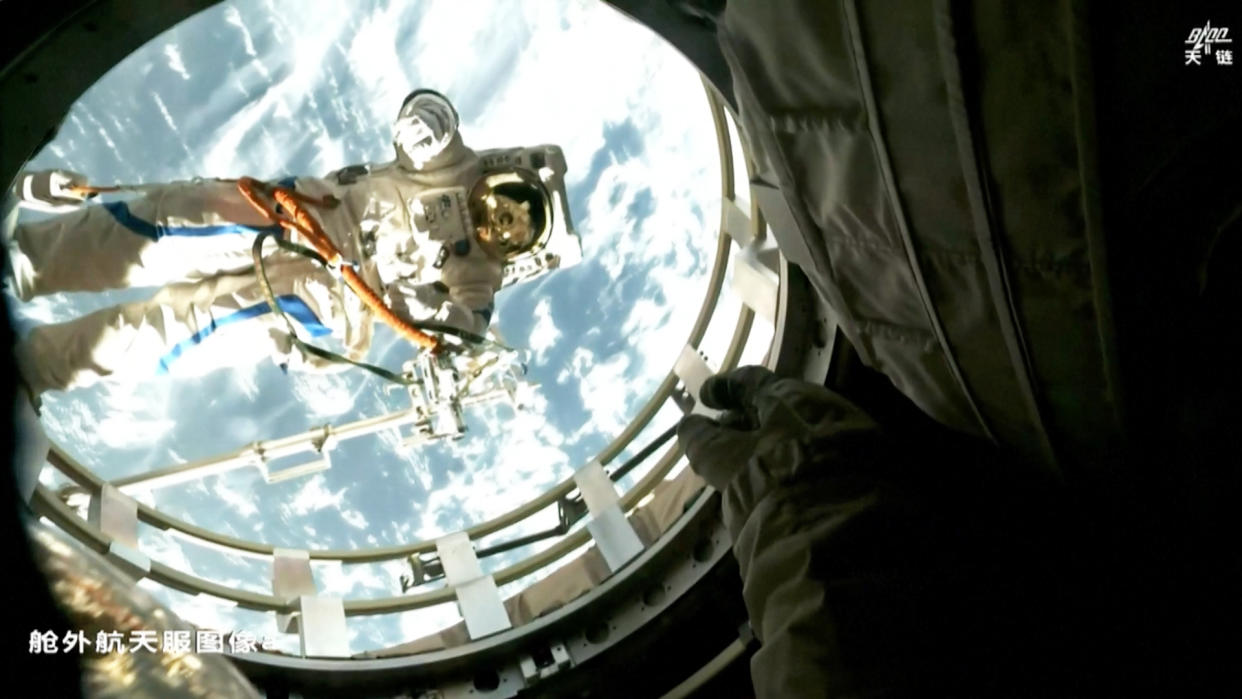  A spacewalking astronaut in a white spacesuit is seen through the open hatch of his spacecraft, with earth's cloudy skies behind him. 