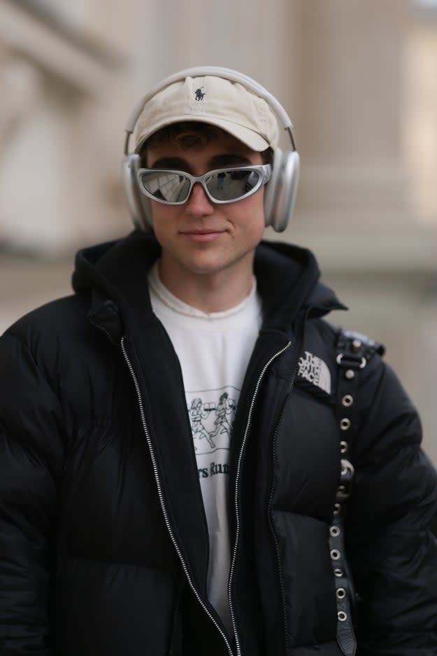 A man wearing headphones, a baseball cap, reflective sunglasses, a white shirt and a dark puffer jacket stands outdoors