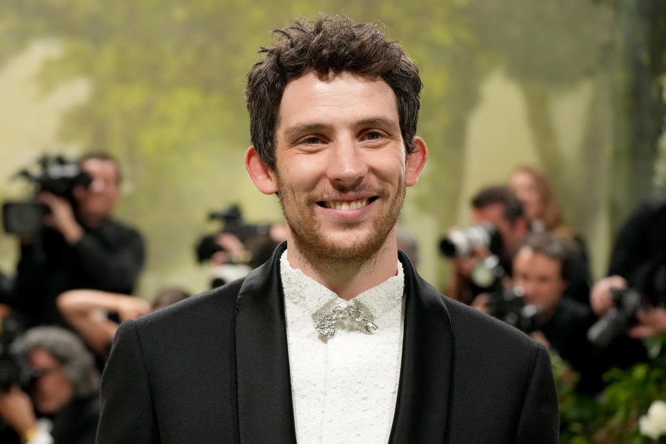 Josh O'Connor smiles on the red carpet, wearing a formal black suit and white shirt with a brooch at the collar. Photographers are in the background