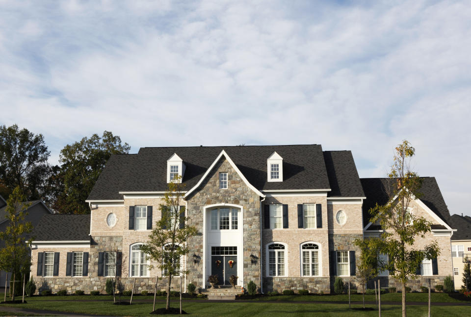 A large, elegant two-story house with brick and stone facade, several windows, and a well-kept lawn. Trees and shrubs are planted in the front yard