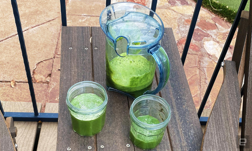A pitcher and two cups sit on a brown wooden table outside. They are filled with green juice made from kale, apples and celery. 