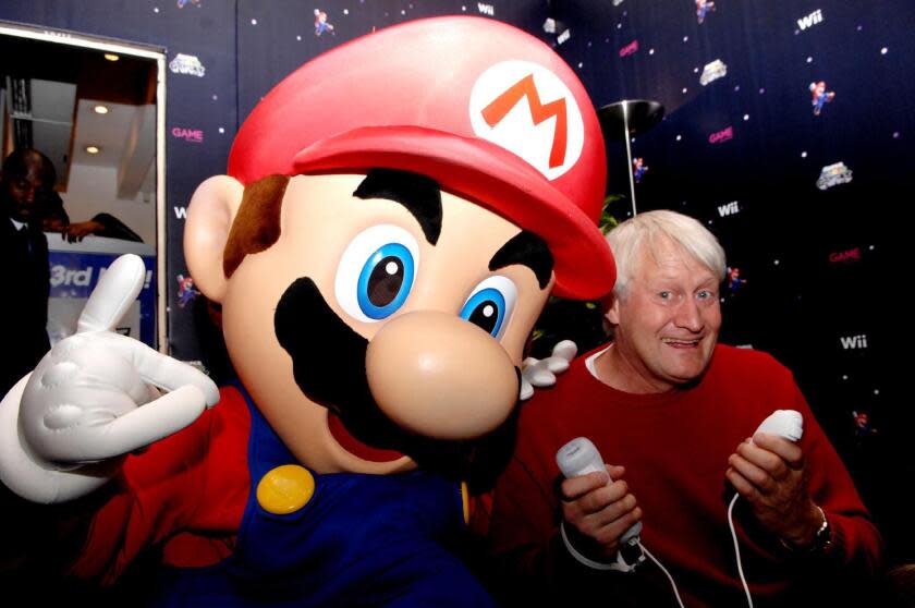 Someone in a Mario costume leaning on Charles Martinet in a red sweater holding Nintendo Wii controllers