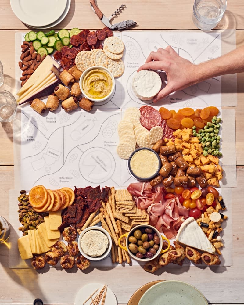 grazing table or board being constructed on 8.5x11 sheets of paper working as templates on a light wood table. There is meat, cheese, crackers, fruit, vegetables, dips, olives, and more. Plates and glasses, toothpicks on the table by the food. Hands are reaching in to place food.