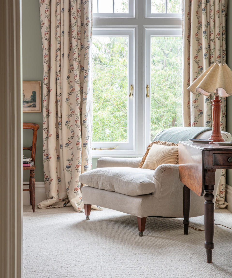 Cozy bedroom with cream wool carpet, relaxed armchair with cushions, dark wood side table with table lamp, large white windows with floral curtains