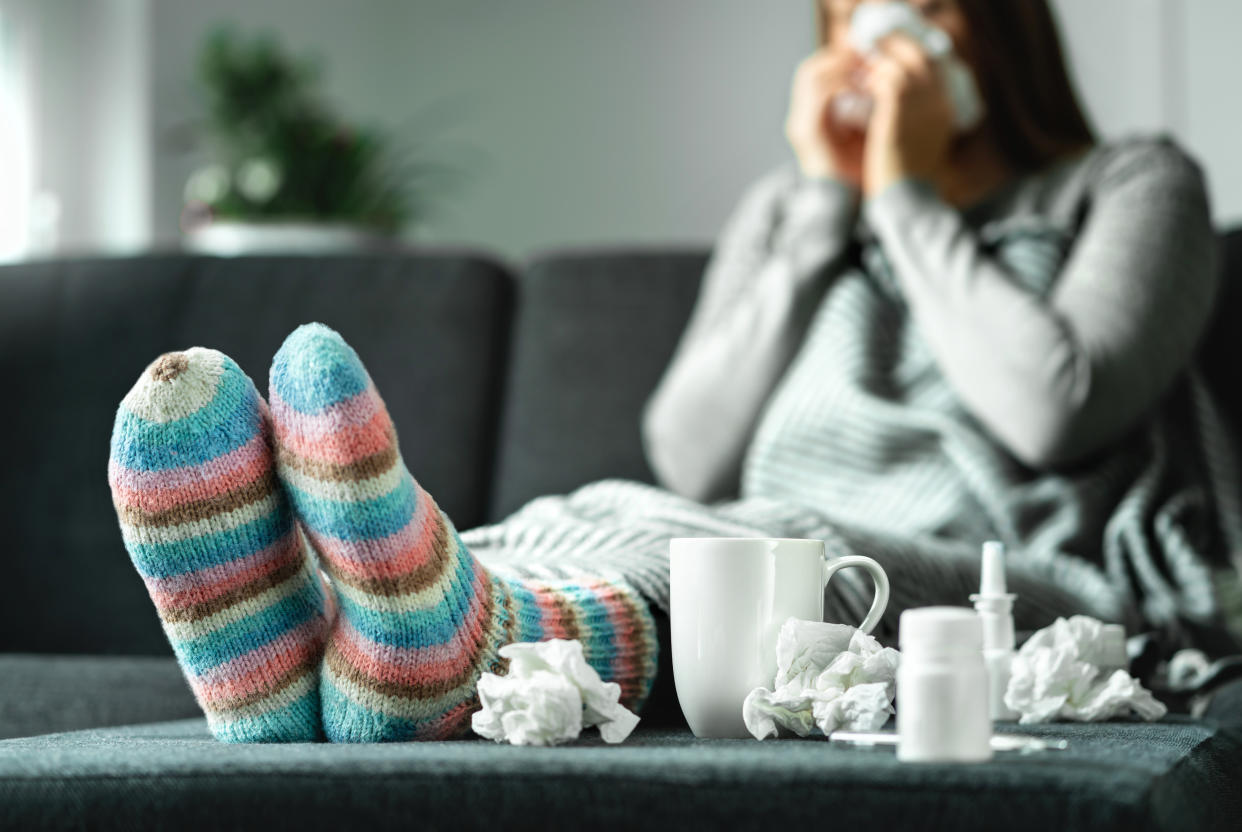 Sick woman with flu, cold, fever and cough sitting on couch at home. Ill person blowing nose and sneezing with tissue and handkerchief. Woolen socks and medicine. Infection in winter. Resting on sofa.