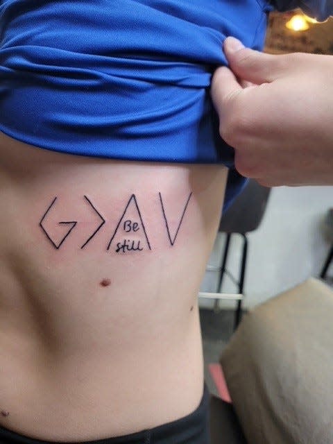 Peyton Kemnitz's tattoo represents the message "God is greater than the highs and lows" using the math "greater than" symbol and arrows.